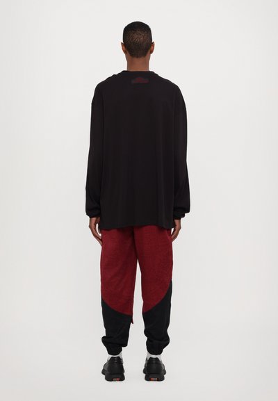 Chemise noire à manches longues, coupe décontractée, associée à un pantalon de jogging à motifs rouges et noirs. Des baskets noires avec des accents orange complètent le look.