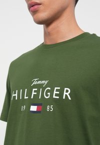 Zelené bavlněné tričko s kulatým výstřihem, výrazným bílým textem a logem s červenými, bílými a modrými akcenty. Design v uvolněném střihu.