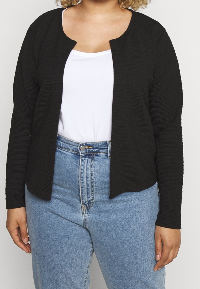 Personne portant un cardigan noir ouvert sur une chemise blanche et un jean bleu taille haute, debout avec les mains détendues sur les côtés.