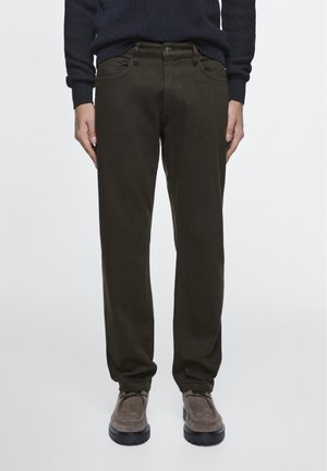 Uomo con pantaloni dritti color oliva scuro, maglione nero e scarpe stringate di camoscio marrone, in piedi davanti a uno sfondo semplice.