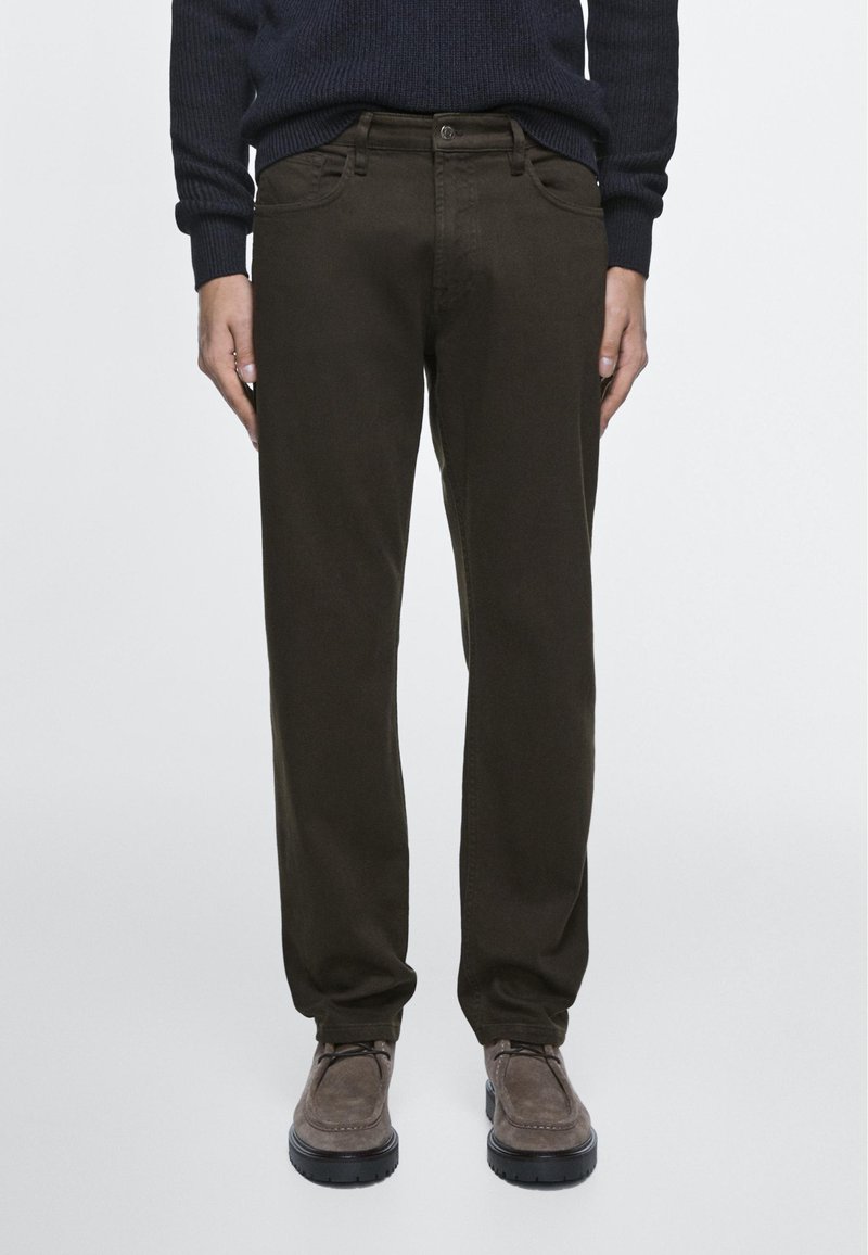 Man die donkere olijfgroene, rechte broek draagt, een zwarte trui en bruine suède veterschoenen, staand tegen een effen achtergrond.