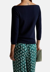 Marineblaue schulterfreie Strickjacke mit geripptem Muster, kombiniert mit grünen gemusterten Hosen und einer kleinen silbernen Clutch.