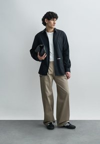 Μαύρο πουκάμισο κουμπωτό πάνω από λευκή κοντομάνικη μπλούζα, καφέ φαρδιά παντελόνια, κρατώντας μαύρο τσαντάκι, με μαύρα παπούτσια που έχουν λευκές ρίγες.