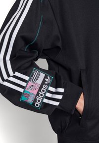Czarna bluza z białym, trzy-paskowym wzorem na rękawach, z kolorową naszywką z logo Adidas na ramieniu; gładka, teksturowana tkanina.