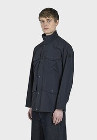 Námořnická modrá bunda vyrobená z lehké tkaniny; má vysoký límec, několik předních kapsiček a uzávěry na patentky.