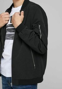 Svart bomberjacka med ribbade muddar och nederkant, framknäppning med dragkedja och en dragkedjeficka på ärmen, bärs över en vit t-shirt.