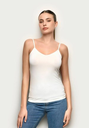 Joven con el cabello recogido, vestida con una camiseta blanca sin mangas y jeans azules, de pie frente a un fondo claro y liso.