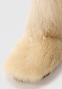 Stivale beige peloso con punta arrotondata, texture morbida e una suola leggermente spessa. Il materiale sembra morbido e caldo.