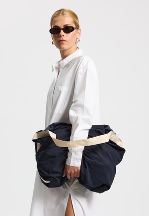 Marineblauer Übergröße-Tasche aus weichem Stoff mit einem cremefarbenen Riemen. Wird von einer Person mit einem weißen Hemd mit Knopfkragen getragen.