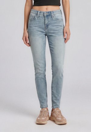 Donna che indossa jeans skinny chiari lavati e scarpe da ginnastica beige, in piedi contro uno sfondo semplice, ritagliata a metà busto e caviglie.