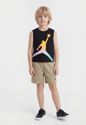 Blond jongetje staat, draagt een zwart mouwloos shirt met kleurrijke basketbalspeler afbeelding, beige korte broek en zwarte sneakers op witte achtergrond.