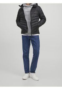 Persona con chaqueta de acolchado negra, sudadera gris, jeans azules y zapatillas blancas, de pie con las manos en los bolsillos de la chaqueta.