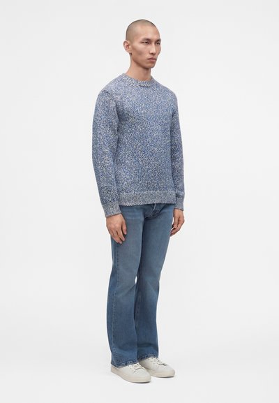 Homme debout portant un pull en maille chiné bleu, un jean bleu décontracté et des baskets blanches sur un fond blanc uni.