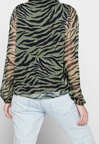 Blusa trasparente nera e verde con motivo a strisce tigre e maniche a sbuffo, abbinata a jeans azzurri chiari. I dettagli includono polsini elastici.