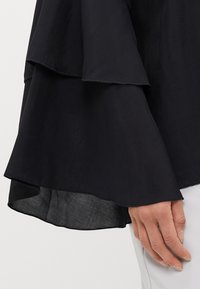 Black top featuring layered ruffle sleeves and a loose fit. The fabric appears lightweight and slightly sheer at the edges.