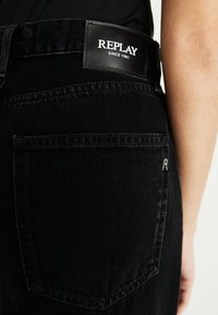 Černé džíny z denimu s koženou nášivkou označenou "REPLAY SINCE 1981." Mají pět kapsový design a decentní detaily v švech.