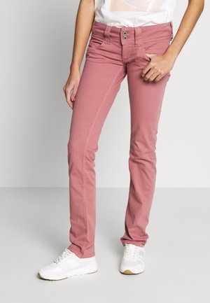 Slim fit dusty pink jeans worn with white sneakers, one hand resting in a pocket, against a plain gray background.