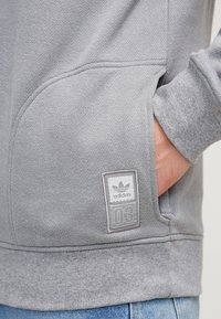 Šedá mikina se strukturovanou tkaninou, s přední kapsou a nášivkou loga Adidas označenou "03" poblíž kapsy.