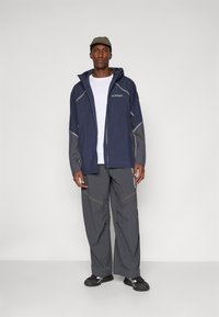 Navy waterproof jacket with grey accents, zipper front, and adjustable hood, paired with loose-fitting grey pants and black shoes.