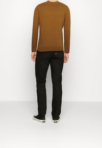Brązowy sweter z długim rękawem wykonany z miękkiego materiału, zestawiony z obcisłymi czarnymi dżinsami oraz czarnymi trampkami z białą podeszwą i detalami.