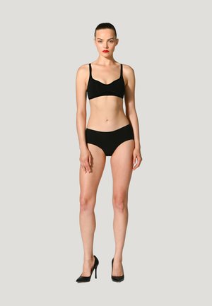 Femme aux cheveux tirés en arrière, portant un bralette noir, un sous-vêtement taille haute assorti et des talons hauts noirs, debout devant un fond clair uni.