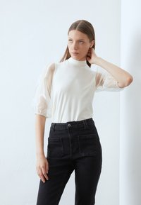 Vit topp med hög krage och genomskinliga puffärmar, matchad med h�ögmidjade svarta jeans med bakfickor. Slät tygtextur.