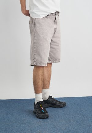 Mand iført lysegrå denimshorts, hvide crew-sokker og sorte sportssko står på et blåt tæppe.