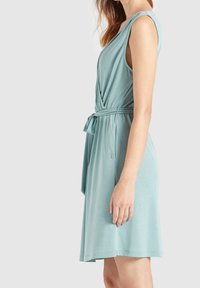 Une robe bleu-vert clair, arrivant au genou, fabriquée en tissu doux avec un corsage torsadé, une taille ceinturée et des manches courtes. Elle comporte un petit accent de logo.