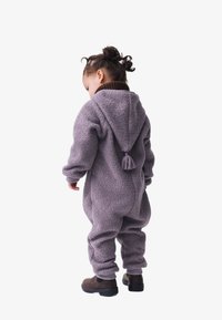 Lila Fleece-Overall mit Kapuze und Quasten-Details, ausgestattet mit umgeschlagenen Ärmel- und Beinabschlüssen, kombiniert mit braunen Stiefeln. Weiche Textur, lockere Passform.