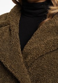Brun tekstureret kunstpelsfrakke med sjalkrave og overlappende design. Sort højhalset bluse under tilføjer kontrast.
