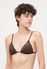 Brunt trekantet bikinioverdel med tynne stropper. Har et samlet design i midten og hvit Nike-logo på den ene siden. Myk, elastisk stoff.