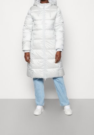 Personne portant un manteau matelassé blanc mi-long avec capuche, un jean bleu clair et des baskets blanches, debout devant un fond uni.