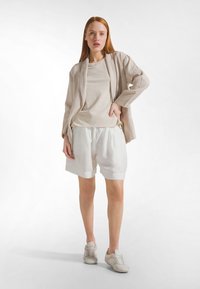 Ljusbeige linnejacka med långa ärmar över en cremefärgad t-shirt, matchad med vita shorts och ljusa sneakers, med avslappnad passform och mjuk textur.