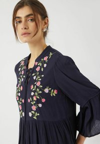 Czarna sukienka w kolorze ciemnoniebieskim z dzwonkowymi rękawami, ozdobiona kwiatowym haftem w odcieniach różu i zieleni. Lekka, teksturowana tkanina z dekoltem w kształcie litery V.