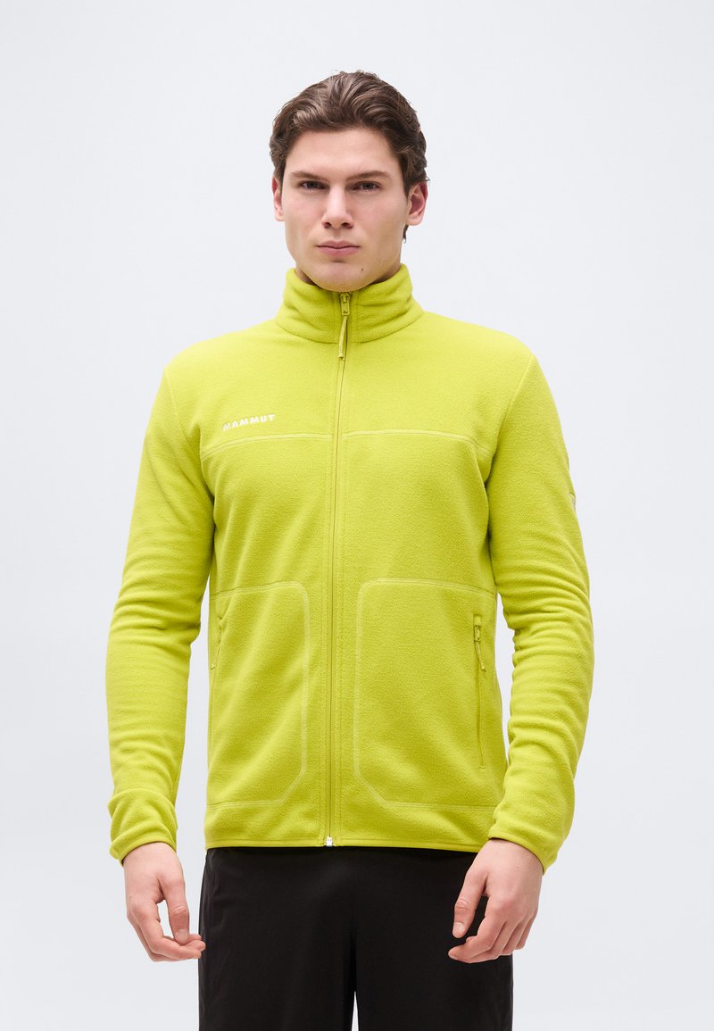 Άνδρας που φορά ένα fleece μπουφάν πλήρους φερμουάρ σε χρώμα λάιμ με ψηλό γιακά και τσέπες με φερμουάρ, στέκεται μπροστά σε ένα απλό λευκό φόντο.