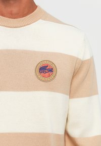 Beige- och vitrandig tröja med ribbad halsringning och ett runt guld- och röd "Tennis Heritage"-märke på bröstet. Mjuk stickad textur.