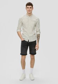 Langärmliges Hemd mit weißen Streifen und Stehkragen, kombiniert mit dunklen Denim-Shorts. Das Model trägt weiße Sneakers und Knöchelsocken.