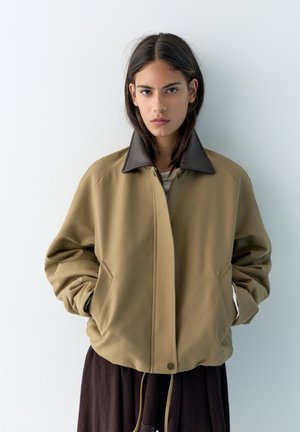 Ung person med langt mørkt hår iført en tanjakke med mørkrabat og hænderne i lommerne, stående mod en ensfarvet hvid baggrund.