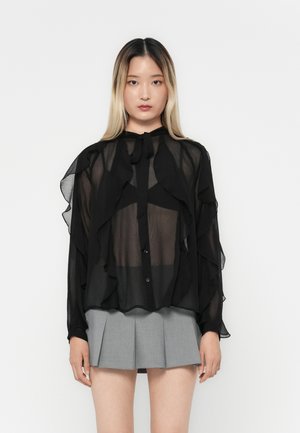 Black sheer blouse with ruffled accents, long sleeves, and a tied neck. Paired with a gray pleated mini skirt.