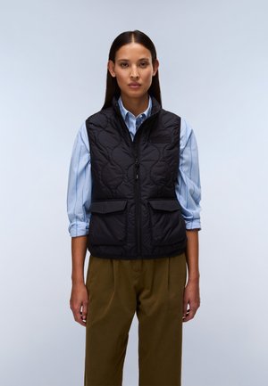 Kvinde med glat mørkt hår iført en sort quiltet vest over en lyseblå, stribet skjorte og olivengrønne bukser, stående mod en ensfarvet baggrund.