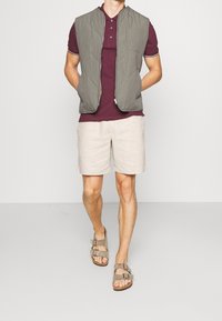 Vadderad grön väst, mörkröd polotröja, ljusbeige shorts och grå sandaler med spännen. Enkel, funktionell design med neutrala färger.