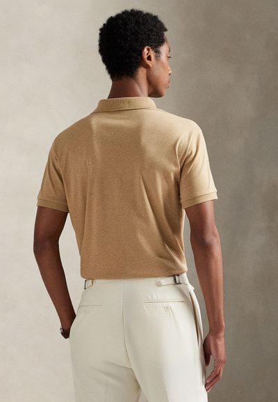 Bărbat cu fața spre spate, purtând un tricou polo bej cu mâneci scurte și pantaloni albi cu reglaje laterale pe un fundal neutru.