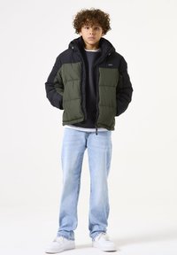 Doudoune olive et noire, avec capuche, fermeture éclair à l'avant et design en panneaux. Associée à un jean bleu clair et des baskets blanches.