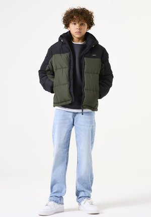 Puffermantel in Olivgrün und Schwarz, mit Kapuze, Reißverschlussfront und Panel-Design. Kombiniert mit hellblauen Jeans und weißen Sneakers.