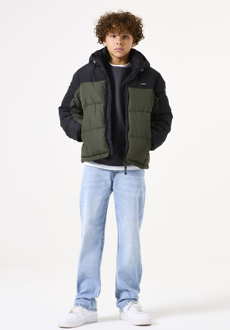 Doudoune olive et noire, avec capuche, fermeture éclair à l'avant et design en panneaux. Associée à un jean bleu clair et des baskets blanches.