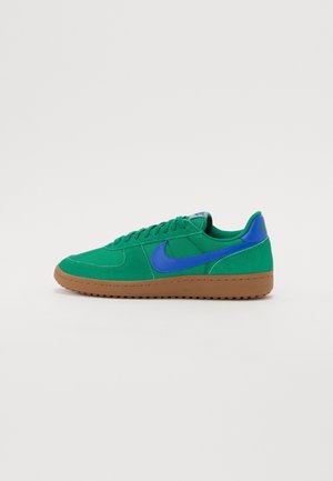 Baskets en daim vert avec le logo Nike en bleu, languette talonnière bleue, semelle en caoutchouc gomme, et lacets verts, présentées de profil sur un fond blanc.