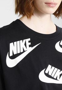 Svart bomull t-shirt med vita Nike-logotyper och swooshs i ett upprepat mönster. Rundad halslinning med korta ärmar. Slät textur.