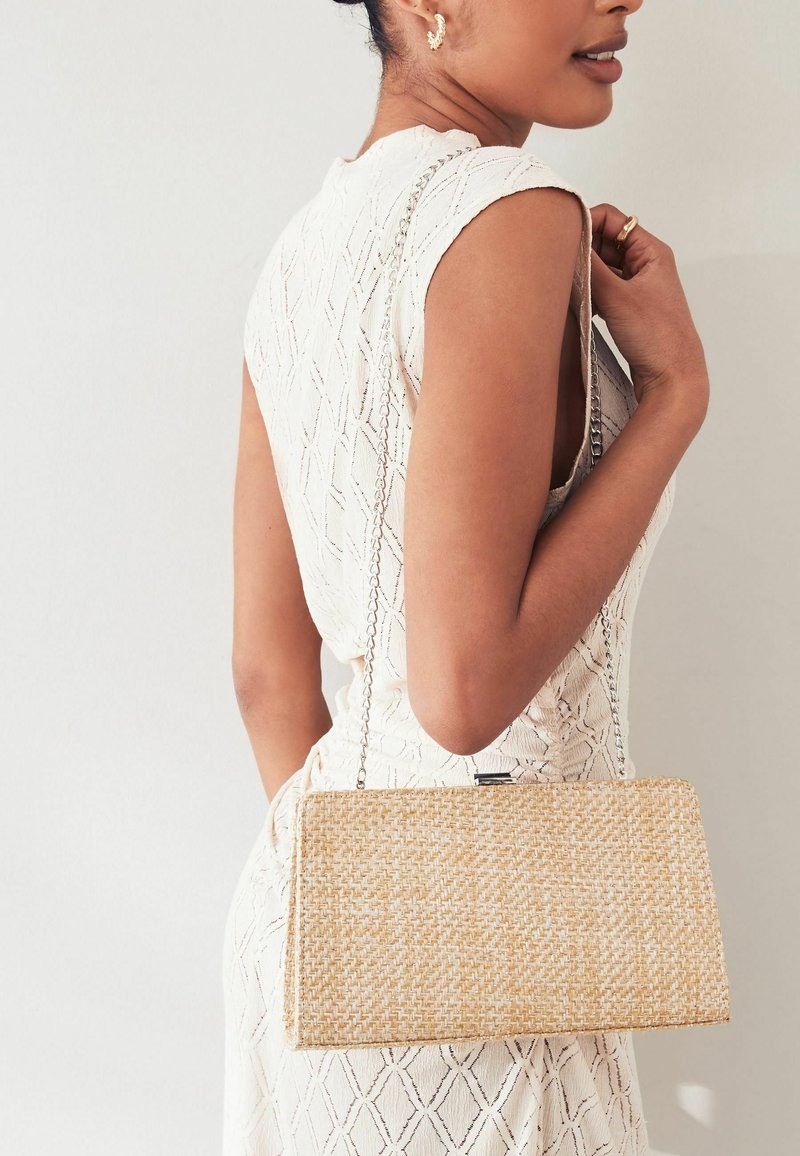 Femme portant une robe blanche sans manches texturée, tenant un sac à main tissé rectangulaire avec une bandoulière en chaîne sur l'épaule.