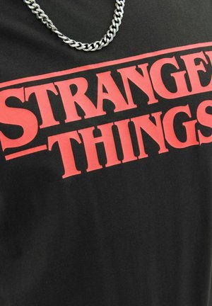Schwarzes T-Shirt mit fettem roten Text, der "STRANGER THINGS" in Großbuchstaben liest, mit einer metallischen Kettenhalskette darüber.