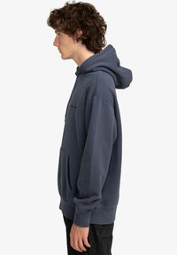 Sudadera azul oscuro hecha de tejido suave, con un bolsillo tipo canguro, puños de canalé y capucha con cordón, mostrada de lado.